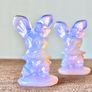 May include: Two opalescent bunny figurines in shades of lavender, blue, and peach. The bunnies have large ears and are standing on a round base. The figurines are displayed on a brown surface with a light blue background.