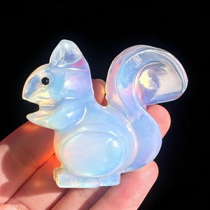 Puede incluir: Una figura de ardilla translúcida e iridiscente con un cuerpo redondeado y una cola tupida. La escultura tiene una superficie lisa y pulida y un ojo negro. La figura se sostiene en una mano sobre un fondo negro.