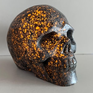 以下が含まれることがあります： 暗い灰色のベースとオレンジ色の斑点模様が特徴的な、装飾用のスカル彫刻。スカルには、眼窩、鼻腔、歯など、詳細な特徴があります。彫刻は、石または同様の素材で作られている可能性があります。