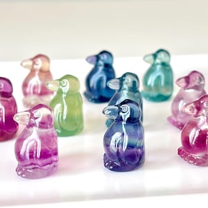 May include: A collection of colorful, carved gemstone penguin figurines. The penguins are in various shades of purple, pink, green, and blue. Each penguin has a small, clear beak and is standing upright. The figurines are displayed on a white surface.