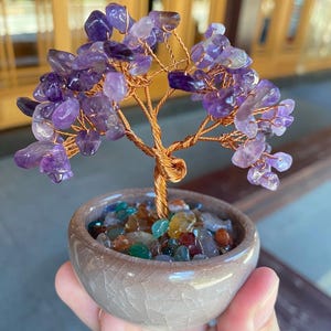 May include: A decorative amethyst gemstone tree with a copper wire trunk and branches. The tree's purple gemstone leaves are clustered, and it sits in a small, round, ceramic pot filled with colorful stones. The pot is approximately 3 inches in diameter.