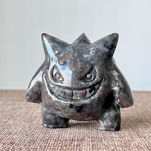 May include: A gray, stone-like figurine of a cartoon character with a wide grin, pointed ears, and menacing eyes. The figure has a textured surface and stands on a brown surface.