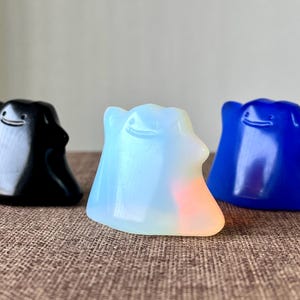 Könnte beinhalten: Drei farbenfrohe, tropfenförmige Figuren mit lächelnden Gesichtern. Eine ist schwarz, eine transluzent weiß mit einem Hauch von Orange und die dritte leuchtend blau. Sie sind auf einer strukturierten, braunen Oberfläche angeordnet.