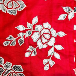 May include: Close-up of a vibrant red fabric with a floral pattern. The design features white, silver-toned, and red embroidered roses and leaves. The fabric has a smooth texture and is suitable for apparel or home decor.