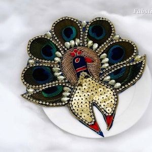 May include: A peacock-shaped embellishment with blue, green, and gold sequins and beads. The peacock's tail feathers are made of green and blue fabric with gold accents. The embellishment is on a white plate.