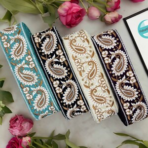 Może przedstawiać: Cztery różne wstążki z tkaniny w wzór paisley. Wstążki są turkusowe, czarne, kremowe i fioletowe. Każda wstążka ma złoty wzór paisley i białe akcenty.