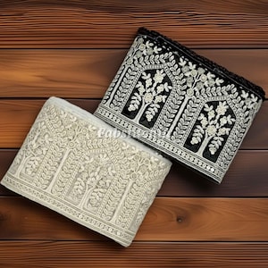 Op de afbeelding: Twee decoratieve stoffen randen, één zwart en één crème, met ingewikkelde bloemenborduursels. De randen hebben een herhalend patroon van gestileerde bomen en bladeren, met een kralenrand. De randen worden op een houten oppervlak weergegeven.