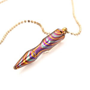May include: A pendant necklace featuring a colorful, cylindrical pendant with a swirling pattern of purple, gold, and teal. The pendant is suspended from a delicate gold-colored ball chain. The background is plain white.
