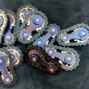 May include: A group of eight metal fidget spinners with black chains and blue and gold accents. The spinners are arranged in a pile on a black surface.