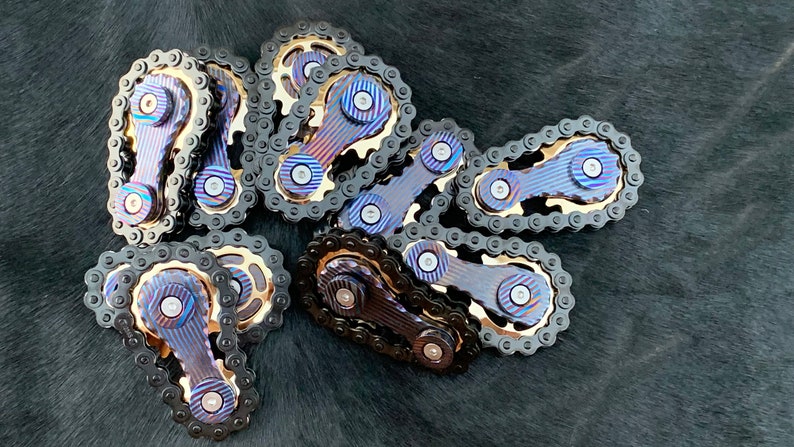 May include: A group of eight metal fidget spinners with black chains and blue and gold accents. The spinners are arranged in a pile on a black surface.