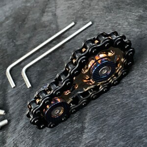 May include: A black chain fidget toy with two spinning gears. The gears are made of metal and have a blue and gold finish. The toy comes with two allen wrenches and two screws.