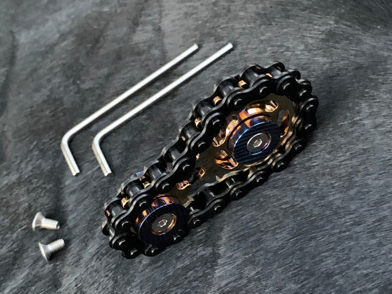 May include: A black chain fidget toy with two spinning gears. The gears are made of metal and have a blue and gold finish. The toy comes with two allen wrenches and two screws.