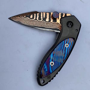 May include: A folding knife with a patterned blade and handle. The blade features a copper and black wave pattern. The handle has a blue, purple, and copper striped design. The knife is open, revealing the blade.