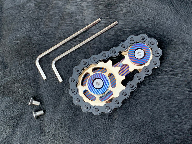 May include: A black chain links to two gold gears with blue and purple accents. The gears have a screw in the center. Two silver Allen wrenches and two silver screws are also included.
