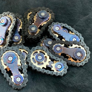 May include: A group of 10 metal fidget toys, each featuring a chain and a gear. The toys are made of a blue and gold metal with a striped pattern.