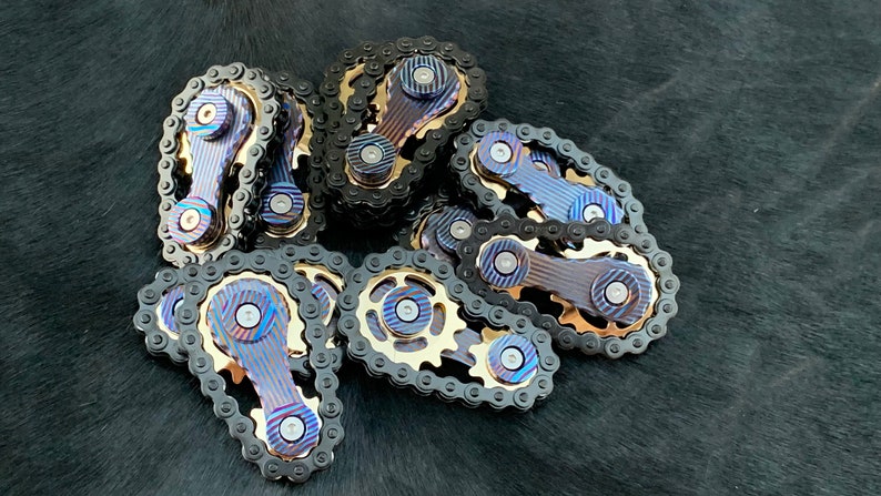 May include: A group of 10 metal fidget toys, each featuring a chain and a gear. The toys are made of a blue and gold metal with a striped pattern.
