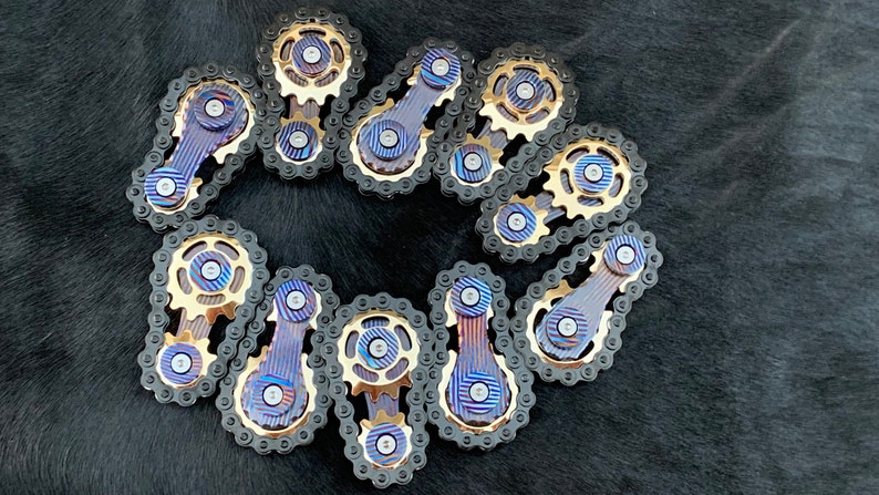 May include: A collection of 12 gold and black metal fidget toys shaped like bicycle chain links. Each link has a small gear and a blue and gold patterned bar.