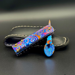May include: A multi-colored pocket knife with a matching spoon, resting on a black leather sheath. The knife and spoon feature a vibrant pattern of blues, oranges, and purples. The sheath has white stitching.