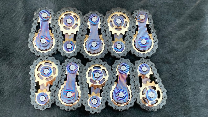 May include: Ten intricately designed metal fidget toys with a blue and gold finish. Each toy features a chain, a gear, and a rotating bar.