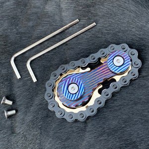 以下が含まれることがあります： 黒のチェーンデザインの青と金のフィジェットスピナー。スピナーには、各リングの中心にネジが付いた、青と金の2つのリングがあります。スピナーには、六角レンチ2本とネジ2本が付属しています。