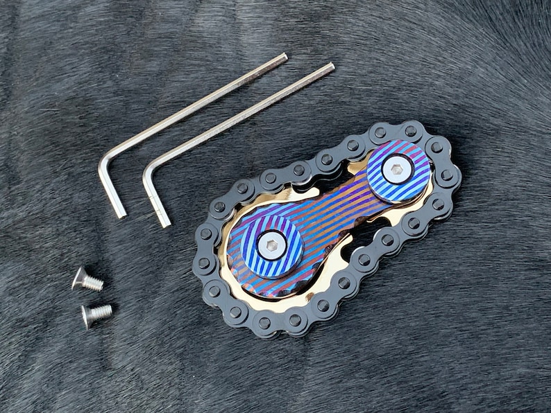 May include: A blue and gold fidget spinner with a black chain design. The spinner has two blue and gold colored rings with a screw in the center of each ring. The spinner comes with two allen wrenches and two screws.