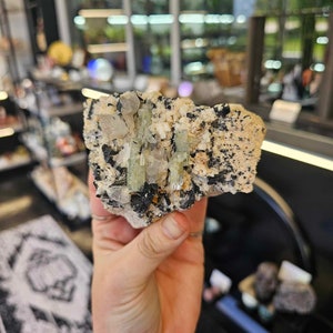 May include: A cluster of green and clear crystals embedded in a rough, gray and black rock. The crystals are elongated and have a glassy appearance.