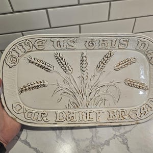 May include: Cream-colored ceramic serving platter with embossed wheat stalks and the words "GIVE US THIS DAY OUR DAILY BREAD" in a decorative font. The platter has a rectangular shape with rounded edges and a textured surface.