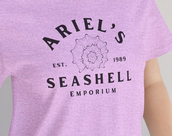 May include: A light purple t-shirt with the text "ARIEL'S SEASHELL EMPORIUM EST. 1989" in black. The design includes a seashell graphic. The shirt is made of a soft material and is suitable for casual wear.
