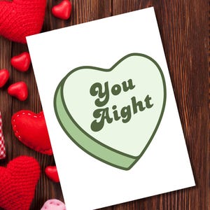 May include: A greeting card with a light green heart shape and the words "You Aight" in dark green. The card is surrounded by red and pink heart-shaped decorations on a wooden background.