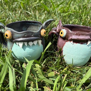 May include: Two ceramic mugs shaped like monsters with protruding eyes, pointed ears, and sharp white teeth. The left mug is dark blue with a light blue belly, while the right mug is dark purple with a light blue belly and two visible fangs.