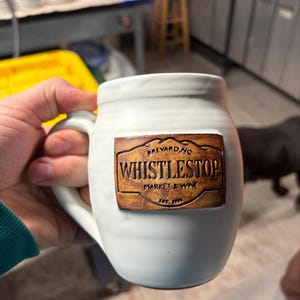 May include: A white ceramic mug with a rounded body and a handle, featuring a rectangular brown clay plaque attached to the front. The plaque is embossed with the text "BREVARD, NC", "WHISTLESTOP", "MARKET & WINE", and "EST. 1995".
