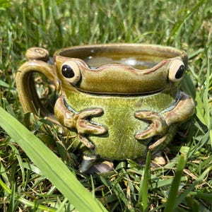May include: A ceramic mug shaped like a frog with a glossy green glaze. The mug features two large, dark, round eyes on the rim, a wide mouth, and two sculpted arms resting on the front. A curved handle is attached to the side of the mug.