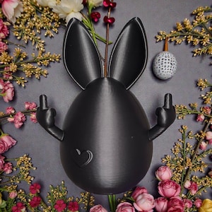 Könnte beinhalten: Eine schwarze, eiförmige Osterfigur mit Hasenohren und Armen, die den Mittelfinger zeigen. Ein Herz ist auf das Ei geätzt. Die Figur ist von rosa, weißen und gelben Blumen und Grün auf grauem Hintergrund umgeben.