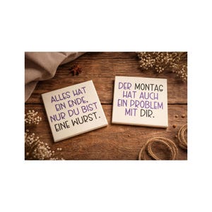 Könnte beinhalten: Zwei weiße quadratische Kacheln mit deutschem Text in lila Tinte auf einer Holzoberfläche. Eine Kachel lautet: "ALLES HAT EIN ENDE. NUR DU BIST EINE WURST." Die andere lautet: "DER MONTAG HAT AUCH EIN PROBLEM MIT DIR."