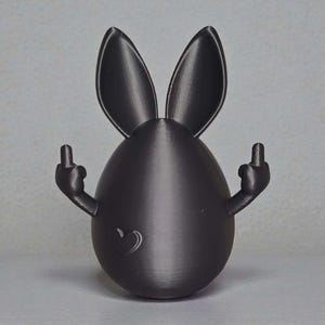 May include: A black, 3D-printed Easter egg figure with bunny ears and arms, displaying the middle finger. A heart is embossed on the egg's surface. The figurine is set against a plain grey background.