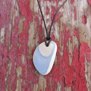 Pode incluir: Um colar com pingente artesanal com uma pedra lisa em forma oval, pintada em azul claro e branco. A pedra está suspensa por um cordão castanho. O fundo é uma superfície de madeira envelhecida com tinta vermelha descascada, dando um aspeto rústico.