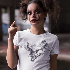 May include: A white t-shirt featuring a black and white graphic print of a skull with antlers. The skull is surrounded by a cloud-like design. The person wearing the shirt has clown makeup on.