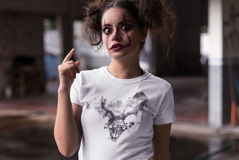 May include: A white t-shirt featuring a black and white graphic print of a skull with antlers. The skull is surrounded by a cloud-like design. The person wearing the shirt has clown makeup on.