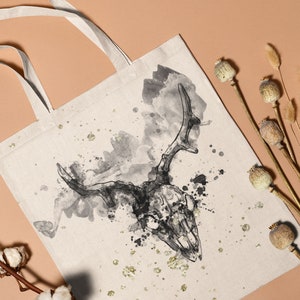 May include: A white canvas tote bag featuring a black and white watercolour illustration of a deer skull with antlers. The background has a splattered watercolour effect.