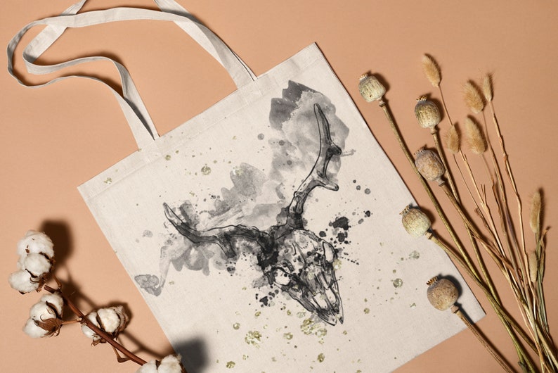 May include: A white canvas tote bag featuring a black and white watercolour illustration of a deer skull with antlers. The background has a splattered watercolour effect.
