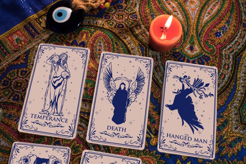 May include: Three tarot cards on a patterned fabric background. The cards are "Temperance", "Death", and "The Hanged Man". Each card features a blue silhouette on a light pink background with a decorative border.