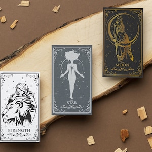 May include: Three tarot cards on a wooden surface. The cards are decorated with gold and silver foil and feature illustrations of a woman with a lion, a woman standing with her arms outstretched, and a woman standing in front of a crescent moon. The cards are labeled "Strength", "The Star", and "The Moon".