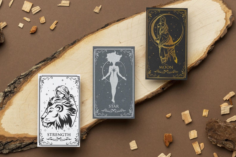 May include: Three tarot cards on a wooden surface. The cards are decorated with gold and silver foil and feature illustrations of a woman with a lion, a woman standing with her arms outstretched, and a woman standing in front of a crescent moon. The cards are labeled "Strength", "The Star", and "The Moon".