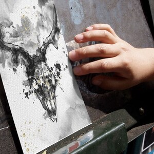 May include: A black and white ink drawing of a deer skull with gold accents on a white background. The drawing is on a piece of paper that is being held by a hand.