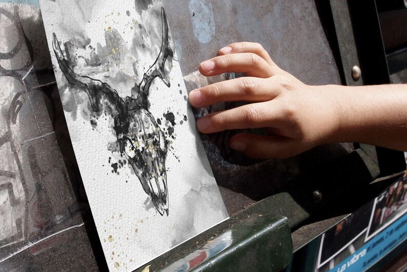 May include: A black and white ink drawing of a deer skull with gold accents on a white background. The drawing is on a piece of paper that is being held by a hand.