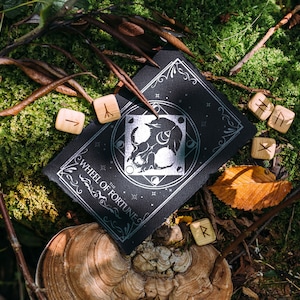 May include: A black card with silver lettering that reads "The Wheel of Fortune" and a silver wolf design. The card is surrounded by wooden runes with Norse symbols.