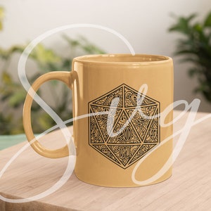 Puede incluir: Una taza de cer&aacute;mica amarilla con un gr&aacute;fico en blanco y negro de un dado de 20 caras. El dado est&aacute; dise&ntilde;ado de forma intrincada con un patr&oacute;n de l&iacute;neas y remolinos.