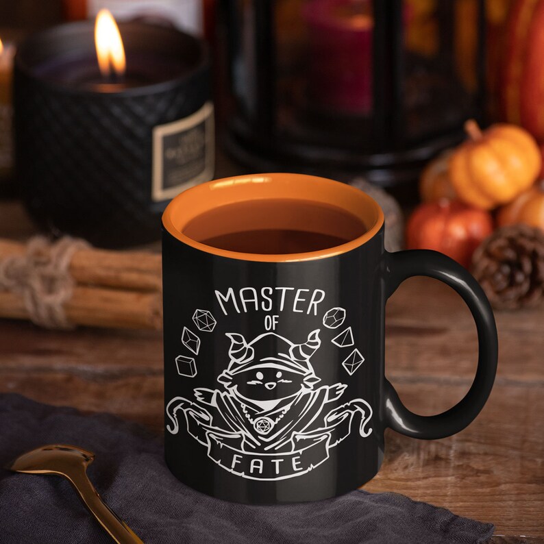 May include: Black coffee mug with an orange rim. The mug features a white illustration of a creature with horns and a scroll that reads "Master of Fate". The creature is surrounded by dice.