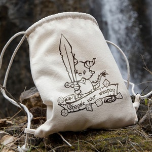 May include: A white drawstring bag with a black and white illustration of a dragon holding a sword. The bag has the text "Choose your weapon" printed on it.