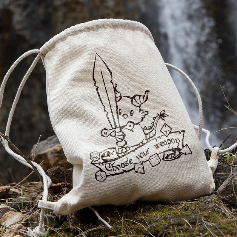May include: A white drawstring bag with a black and white illustration of a dragon holding a sword. The bag has the text "Choose your weapon" printed on it.